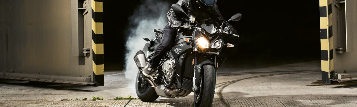 A person in all black protective gear on a BMW® S 1000 R motorcycle screeching out of a hangar.