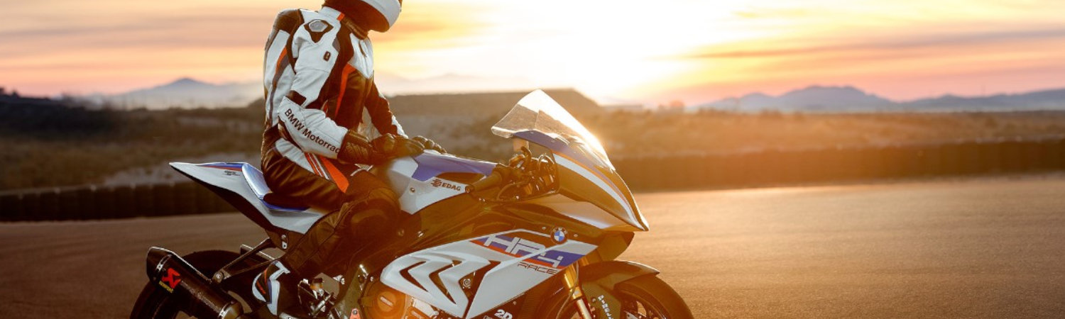 Person in black and white protective gear siting on a BMW HP4 RACE on a track at sunset.